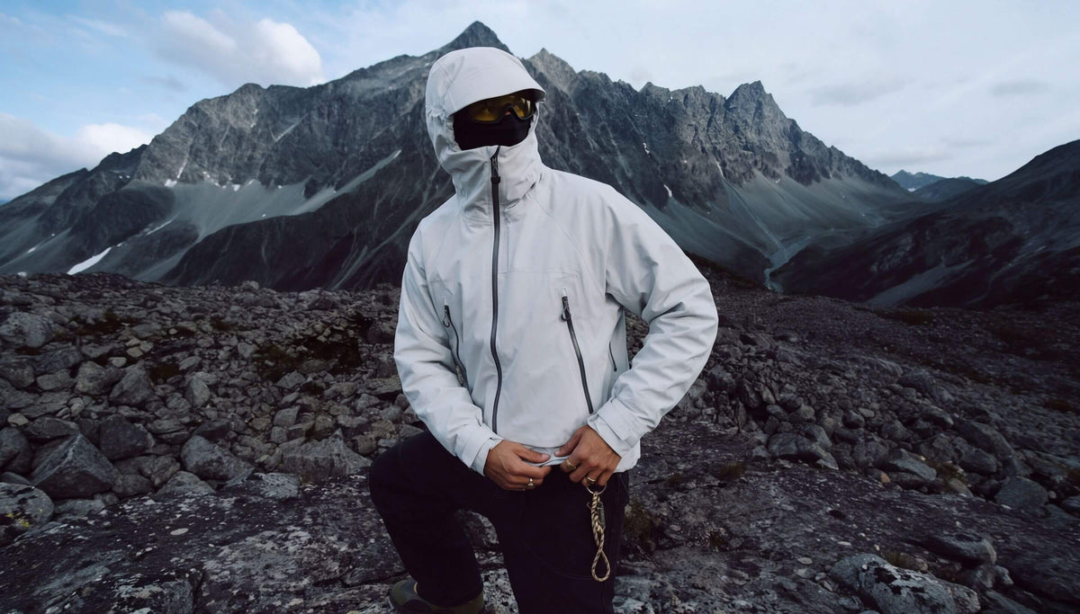 A person wearing a light gray hooded technical jacket and black pants stands on a rocky mountain slope with jagged peaks and a cloudy sky in the background.
