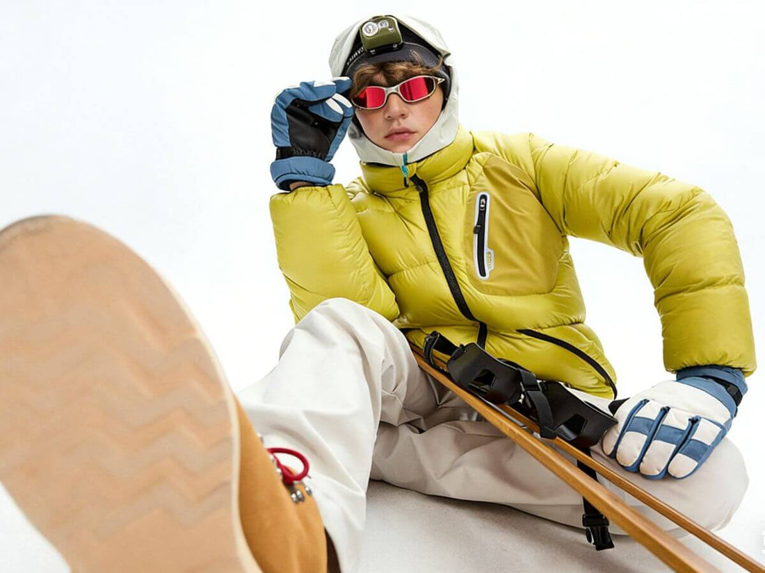A skier wearing a bright yellow puffer jacket, beige ski pants, blue and white gloves, red-tinted goggles, and a white helmet, posed against a clean white background with ski poles and a boot sole in the foreground.