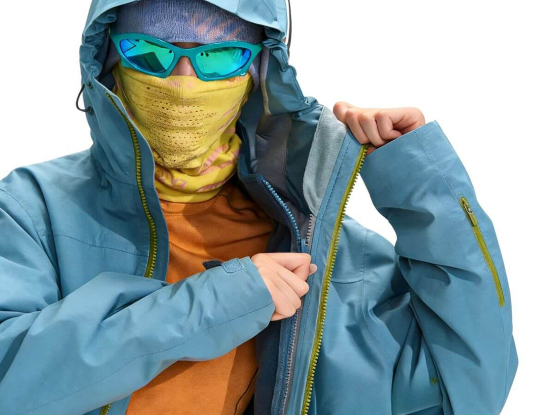 A person wearing a light blue hooded waterproof jacket, a yellow face mask, blue mirrored sunglasses, and an orange shirt, shown against a plain white background while zipping the jacket.