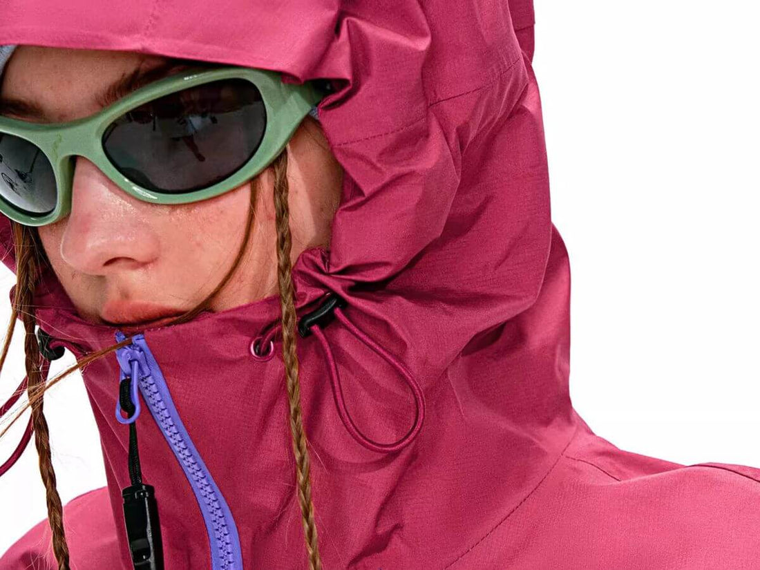 Close-up of a person wearing a bright pink hooded jacket with a purple zipper and green sunglasses against a white background.