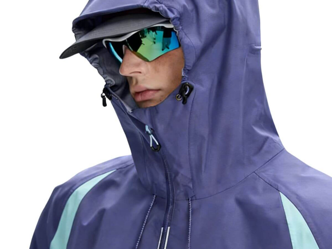 A close-up of a person wearing a hooded rain jacket in a muted periwinkle or lavender-purple color with light aqua side panels, along with a black visor and mirrored sunglasses, against a plain white background.