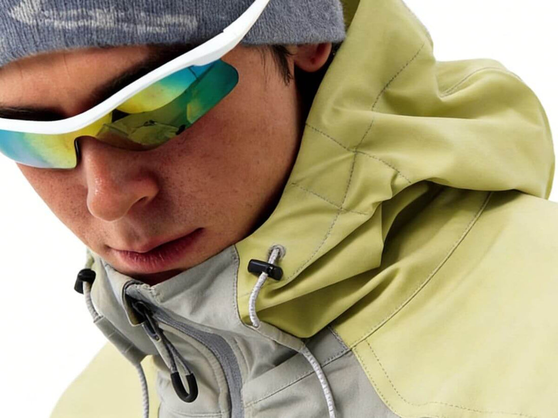 Close-up of a person wearing a light olive green hooded jacket with a gray inner layer, a gray beanie, and mirrored wraparound sunglasses against a white background.